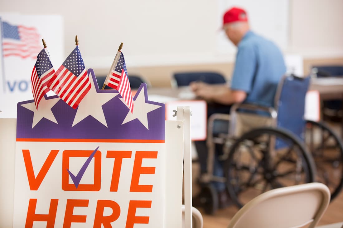 "Vote Here" sign and a man in wheelchair filling out a ballot