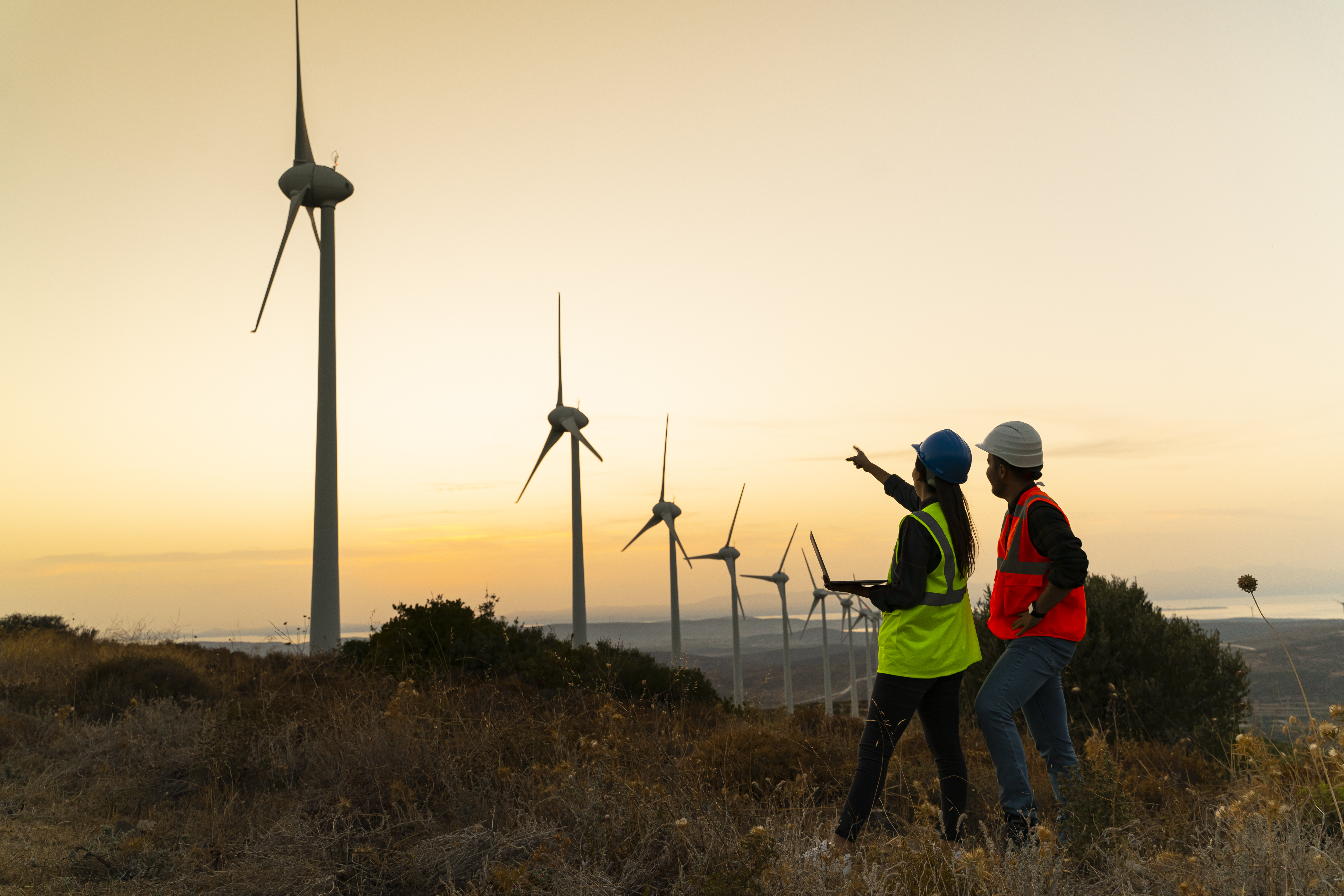 Two workers on iPads examining a line of wind turbines off in the distance.